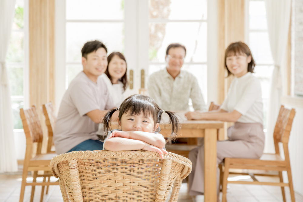 A three generation family sits at the kitchen table while young girl smiles at camera.